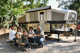 A family at a picnic table beside a tent-trailer under the trees